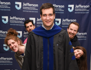 PhD Graduate Standing in Front of Four People Peeking Out from Behind Him